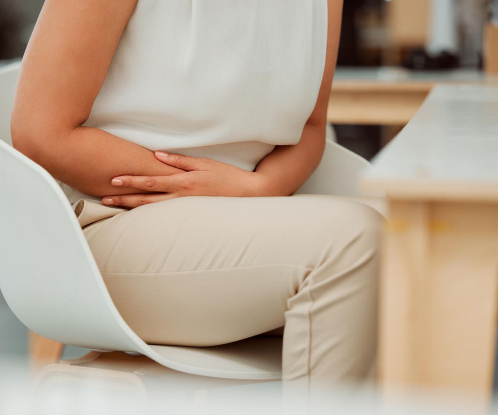 Kobieta w beżowych spodniach i jasnej bluzce siedzi na białym krześle, trzymając dłonie splecione na brzuchu, co symbolizuje dyskomfort związany z pęcherzem. Przeczytaj na Poradnik Zdrowie, jak pozycja siedząca wpływa na zdrowie pęcherza.