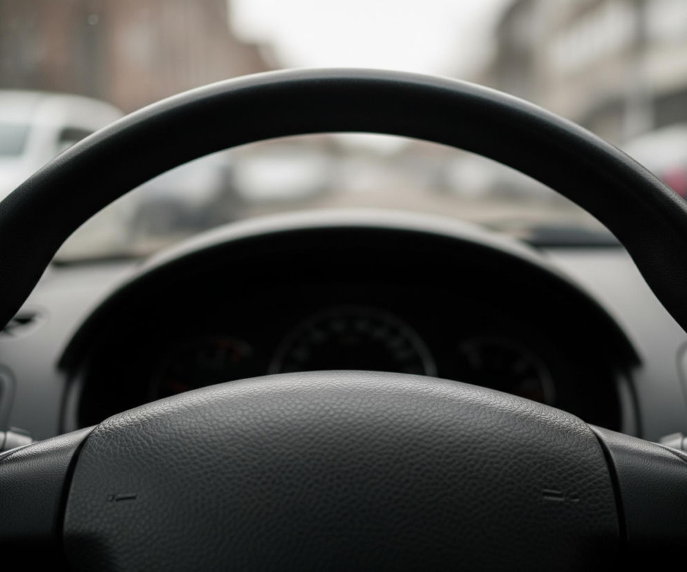 Centralny element zdjęcia to czarna kierownica samochodu, dominująca w kadrze. Jej teksturowana powierzchnia jest matowa i wygląda na skórzaną lub gumową, z widocznymi, drobnymi nierównościami materiału. Za kierownicą, w górnej części kadru, przez przednią szybę widać niewyraźne, rozmyte tło miejskiego krajobrazu z zarysami budynków i innych samochodów w odcieniach szarości i bieli, co sugeruje pochmurny dzień lub deszcz. Poniżej widoczny jest fragment deski rozdzielczej z nieostrymi wskaźnikami.
