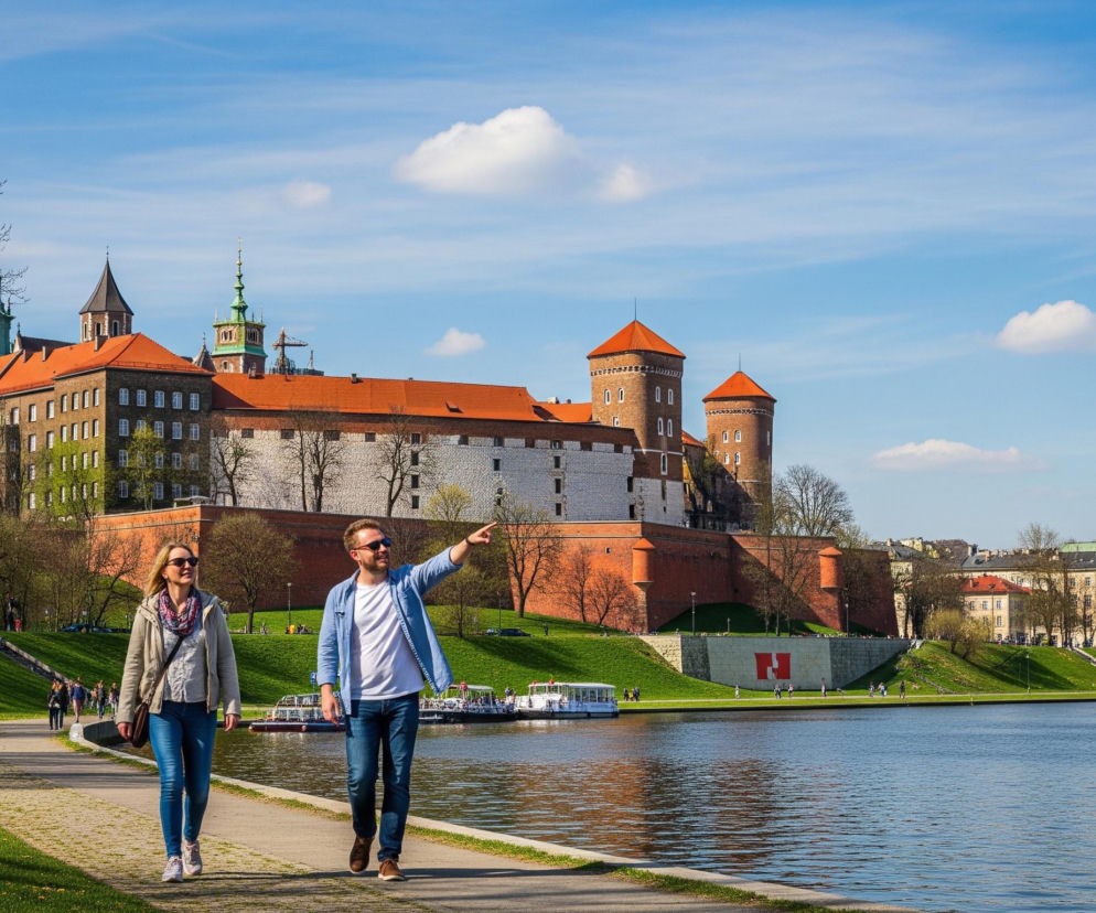 Dwoje ludzi idzie brukowaną ścieżką wzdłuż rzeki, na pierwszym planie po prawej. Mężczyzna w niebieskiej koszuli i okularach przeciwsłonecznych, trzymając rękę wyciągniętą w kierunku oddalonego budynku, rozmawia z kobietą w jasnej kurtce i szaliku. Po lewej stronie ścieżki rozciąga się zielony trawnik z widocznymi żółtymi kwiatami. Na drugim planie, za rzeką, dominuje duży kompleks zamkowy z czerwonymi dachami i kilkoma wieżami, otoczony zielonymi drzewami i ceglanym murem. W oddali, po prawej stronie, widać panoramę miasta z wieloma budynkami i kolejnymi drzewami, a na horyzoncie rozciąga się jasne, błękitne niebo z kilkoma chmurami.