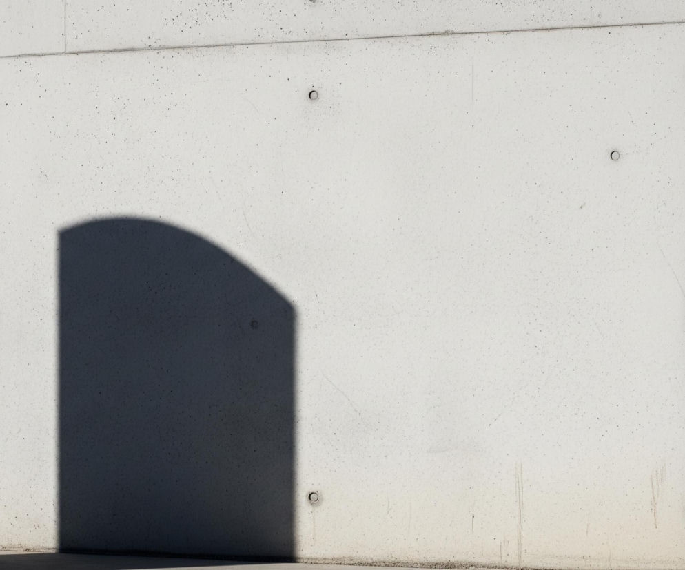 Centralną częścią obrazu jest jasnoszary betonowy mur, podzielony na trzy segmenty pionowymi i poziomymi liniami. Na lewej części muru, w dolnej połowie, widoczny jest ciemny, półkolisty cień przypominający kształt łuku lub wnęki, rzucany przez niewidoczną konstrukcję. Na powierzchni betonu rozmieszczone są liczne małe okrągłe otwory, a także drobne zabrudzenia i ślady. Dół ściany łączy się z poziomym podłożem, które również jest jasnoszare i częściowo zacienione.