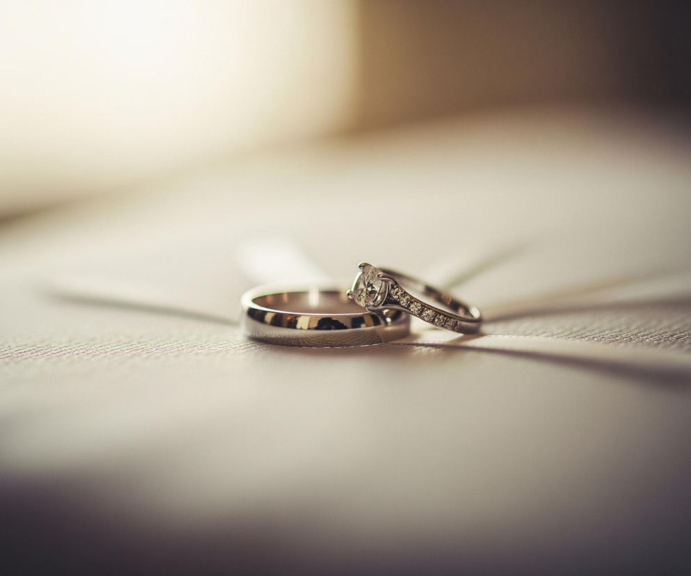 Dwa srebrne pierścionki leżą na jasnej, teksturowanej powierzchni. Większy pierścionek, o gładkiej powierzchni, jest położony płasko, podczas gdy mniejszy, z osadzonymi w nim kamieniami i centralnym, większym diamentem, jest lekko przechylony, opierając się o większy pierścionek. Całość jest sfotografowana z płytką głębią ostrości, co sprawia, że tło jest rozmyte, tworząc delikatne przejście od jasnego, niemal białego światła w górnej części do ciemniejszych, brązowawych odcieni w dolnej części obrazu. Na powierzchni widać subtelne cienie rzucane przez pierścionki.