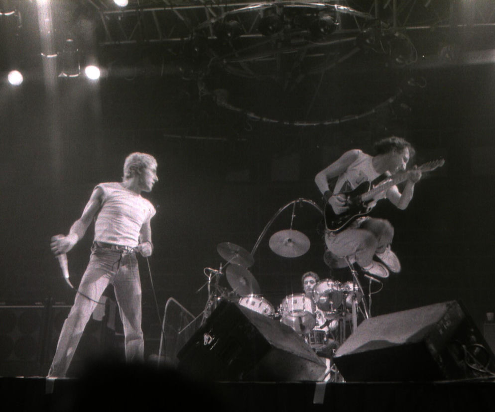 Czarno-białe zdjęcie z koncertu The Who, na którym wokalista Roger Daltrey stoi z mikrofonem, a gitarzysta Pete Townshend wyskakuje w powietrze z gitarą. W tle widoczna perkusja. Artykuł o ich legendarnej bójce i historii zespołu znajdziesz na naszym portalu.