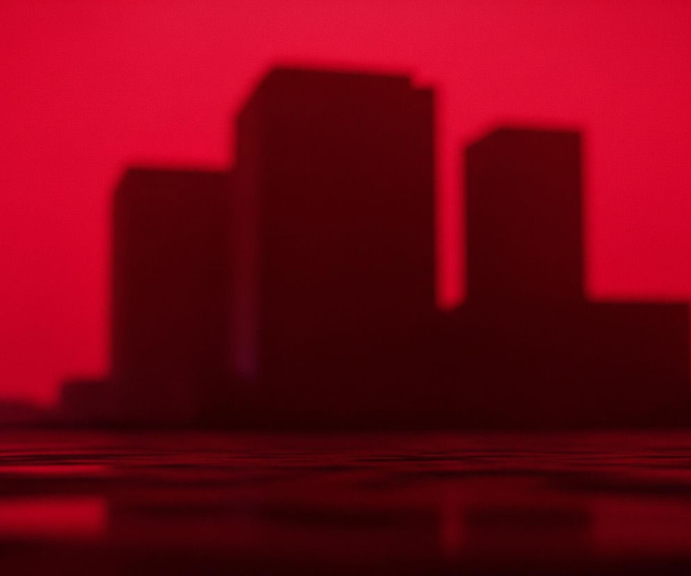 Na pierwszym planie, w lewym dolnym rogu, rozmyte, okrągłe światła w kolorach niebieskim i czerwonym odbijają się na lśniącej, ciemnoczerwonej powierzchni. W tle, na intensywnie czerwonym niebie, widać trzy ciemne, zamazane sylwetki budynków o prostych kształtach, tworzące panoramę miasta. Całość jest skąpana w dominującym odcieniu czerwieni, z odcieniami od jaskrawoczerwonego nieba po głęboki burgund na powierzchni odbijającej światło.