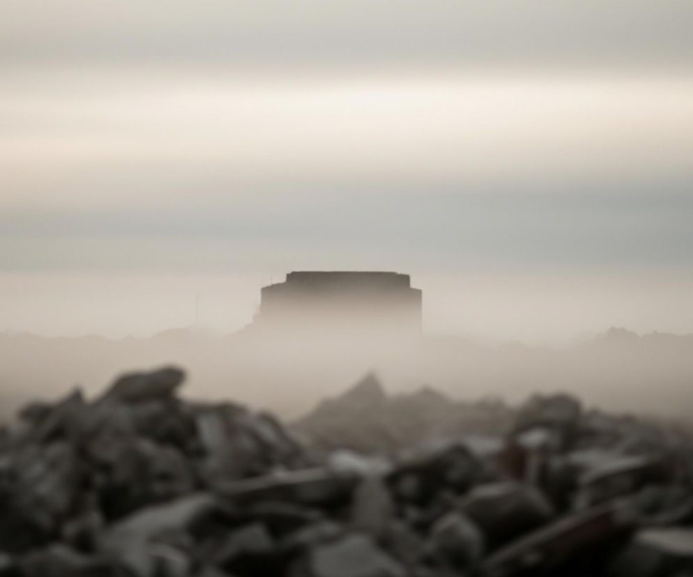 Na pierwszym planie, u dołu obrazu, widoczny jest rozmyty stos gruzu i kamieni w odcieniach szarości i brązu. Za nim rozciąga się mglista, beżowo-szara przestrzeń, w której na horyzoncie rysuje się niewyraźna, ciemna sylwetka budynku o prostej, prostokątnej bryle, z płaskim dachem i delikatnym wgłębieniem w środkowej części. Niebo powyżej jest jasne, w górnej części przechodzi w ciemniejszy, szary odcień, z delikatnym, jaśniejszym pasmem w środku, a cała scena spowita jest w jednolitą, bladą mgłę lub światło, nadając obrazowi monochromatyczny charakter.