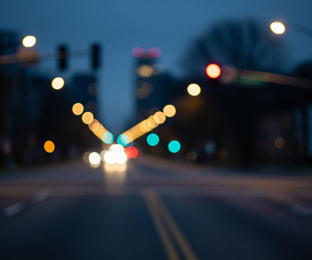 Asfaltowa ulica w kolorze granatowym rozciąga się od dołu kadru, z dwoma żółtymi, rozmytymi liniami biegnącymi przez środek. Po obu stronach drogi widoczne są rozmyte światła w odcieniach żółci, pomarańczy, bieli, czerwieni i turkusu, ułożone w rzędy. W oddali, po lewej i prawej stronie, widać niewyraźne zarysy ciemnych drzew i budynków, a nad nimi ciemnogranatowe niebo.