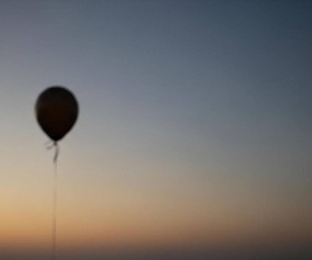 Rozmyty, ciemny balon unosi się na tle gradientowego nieba. Lewa dolna część nieba ma ciepłe, pomarańczowo-żółte odcienie, które przechodzą w chłodniejsze, błękitno-szare barwy w górnej prawej części. Cienki sznurek, również rozmyty, zwisa z balonu w dół obrazu.