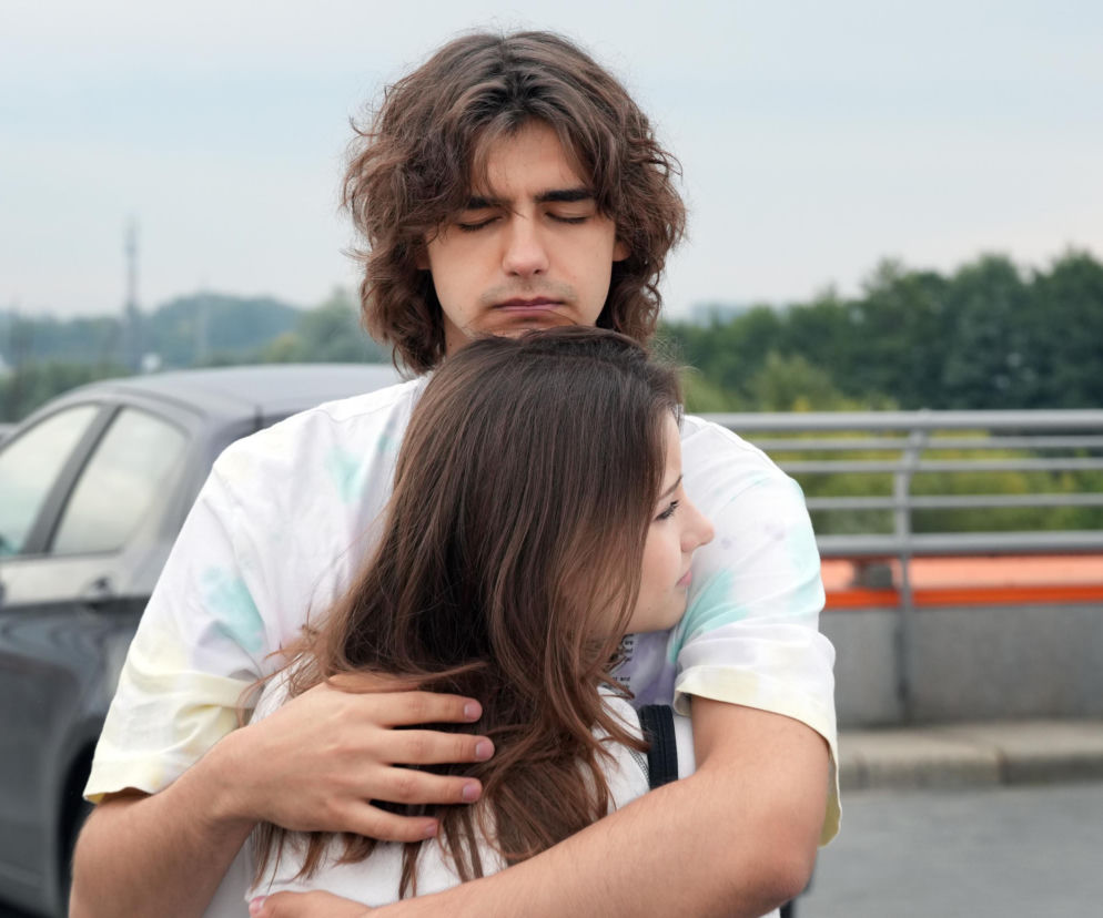 Zasmucony młody mężczyzna, Romeo (Jacek Tyszkiewicz), o długich, kręconych włosach, z zamkniętymi oczami, obejmuje Ewę (Gabriela Ziembicka) na tle szarego samochodu. Zdjęcie przedstawia bolesne pożegnanie pary, która musi się rozstać, co podkreśla emocje w odcinku serialu Barwy szczęścia dostępnym na Super Seriale.