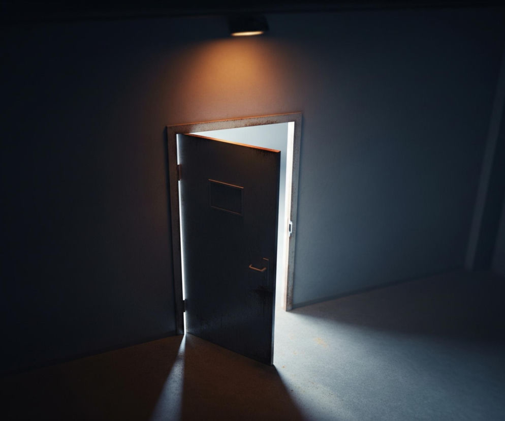 Ciemne, niebieskoszare pomieszczenie jest częściowo oświetlone przez uchylone metalowe drzwi i pojedynczą lampę sufitową. Intensywne, białe światło wydobywa się ze szczeliny drzwi, tworząc ostry, stożkowy promień na podłodze. Lampa rzuca ciepłe, pomarańczowe światło na ścianę nad drzwiami, kontrastując z chłodną tonacją reszty pomieszczenia. Na drzwiach widoczny jest mały, prostokątny otwór.