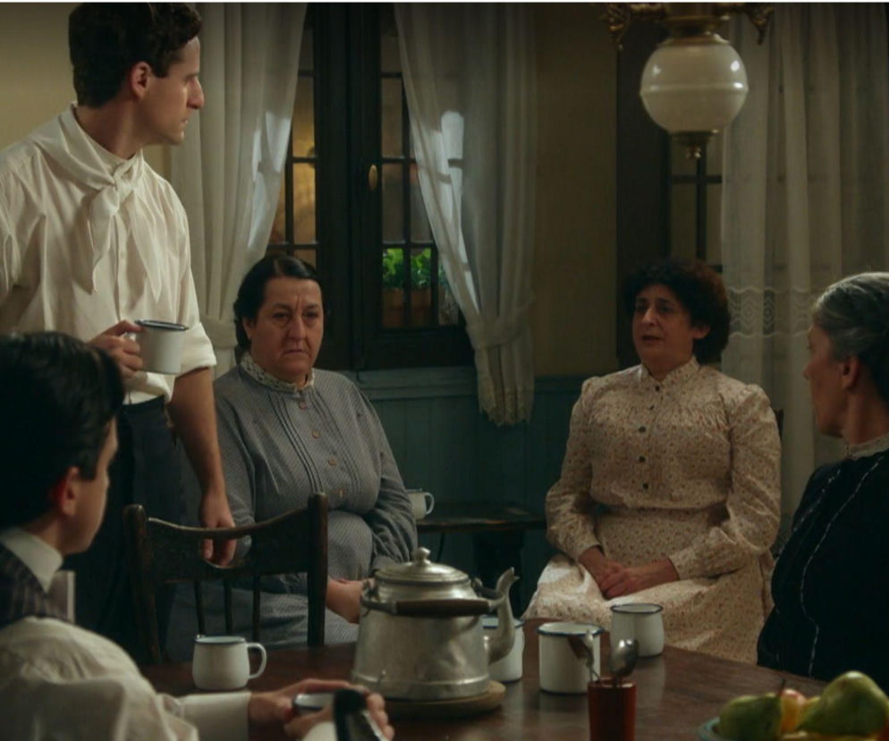 Scena z serialu „La Promesa”, gdzie mężczyzna w jasnej koszuli trzyma kubek, stojąc przy stole, przy którym siedzą cztery kobiety. Na stole widoczne są filiżanki, dzbanek i owoce. Zdjęcie ilustruje codzienne życie bohaterów, o którym możesz przeczytać na portalu Super Seriale.
