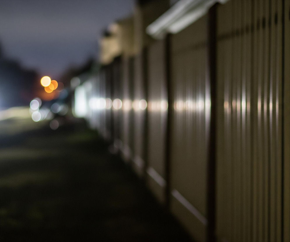 Prawa strona zdjęcia przedstawia długi, beżowy płot z pionowych paneli, który rozciąga się w głąb obrazu, stopniowo tracąc ostrość. Na płocie widać rzędy jasnych, rozmytych punktów świetlnych, pochodzących od latarni lub innych źródeł światła. Lewa strona ukazuje ciemną ulicę lub drogę, na której widoczne są dwa jasne, rozmyte reflektory nadjeżdżającego samochodu oraz kilka pomarańczowych i białych świateł w tle. Całość jest nocnym krajobrazem miejskim lub podmiejskim, z rozmazanymi światłami tworzącymi efekt bokeh.