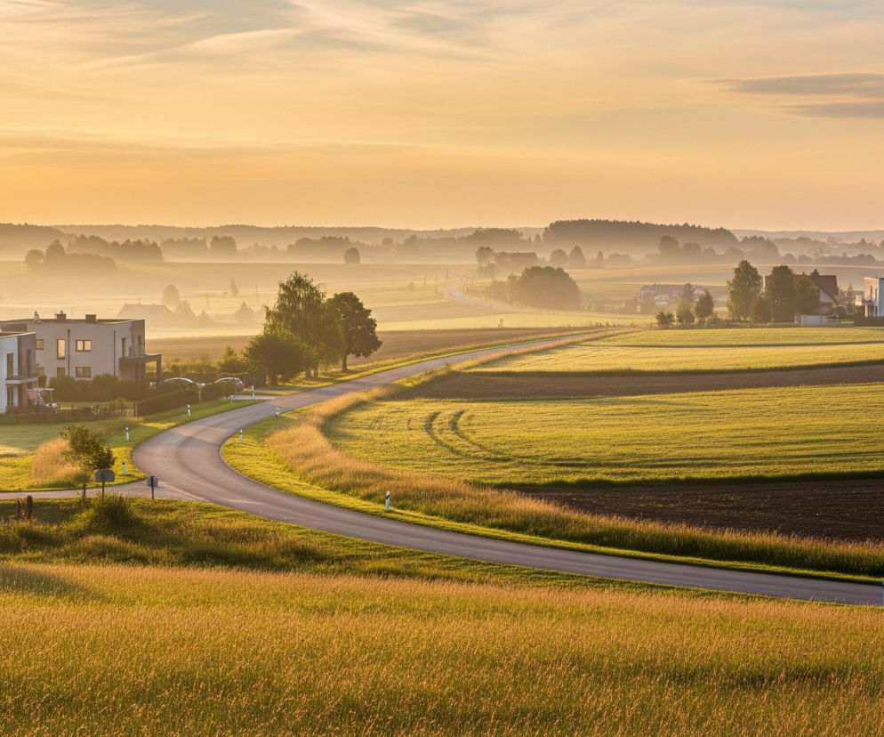 Krzywa droga asfaltowa przecina krajobraz, wijąc się między jasnozielonymi i brązowymi polami uprawnymi. Po lewej stronie drogi widoczne są dwa nowoczesne domy z białymi elewacjami i ciemnymi dachami, otoczone drzewami i krzewami. W tle rozciąga się mglisty krajobraz z lasami i odległymi zabudowaniami, wszystko skąpane w ciepłym, pomarańczowo-żółtym świetle wschodzącego lub zachodzącego słońca. Niebo na górze jest jasne, przechodzące w błękit, z delikatnymi, poziomymi obłokami.