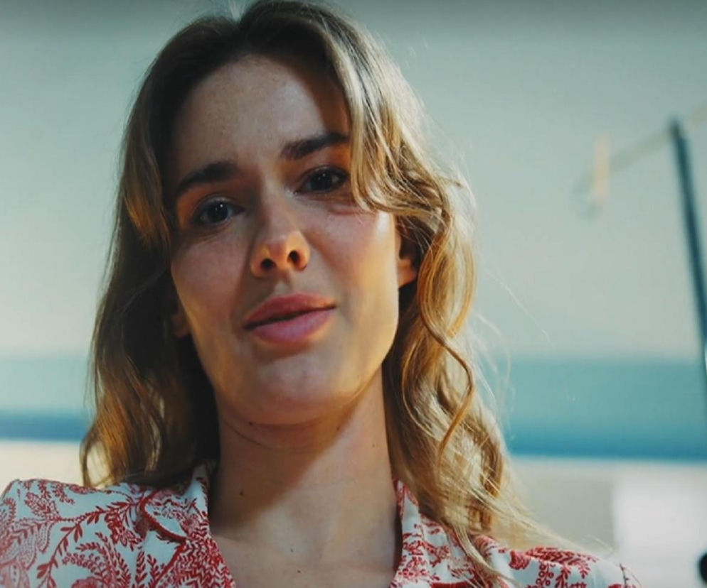 Kobieta o jasnych, falowanych włosach, ubrana w piżamę w czerwono-biały wzór, patrzy w dół z zaniepokojeniem. Tło sugeruje wnętrze szpitala, widoczny statyw medyczny. Scena z serialu M jak miłość, gdzie Franka nagrywa pożegnalne filmiki dla Pawła, co jest dostępne na Super Seriale.