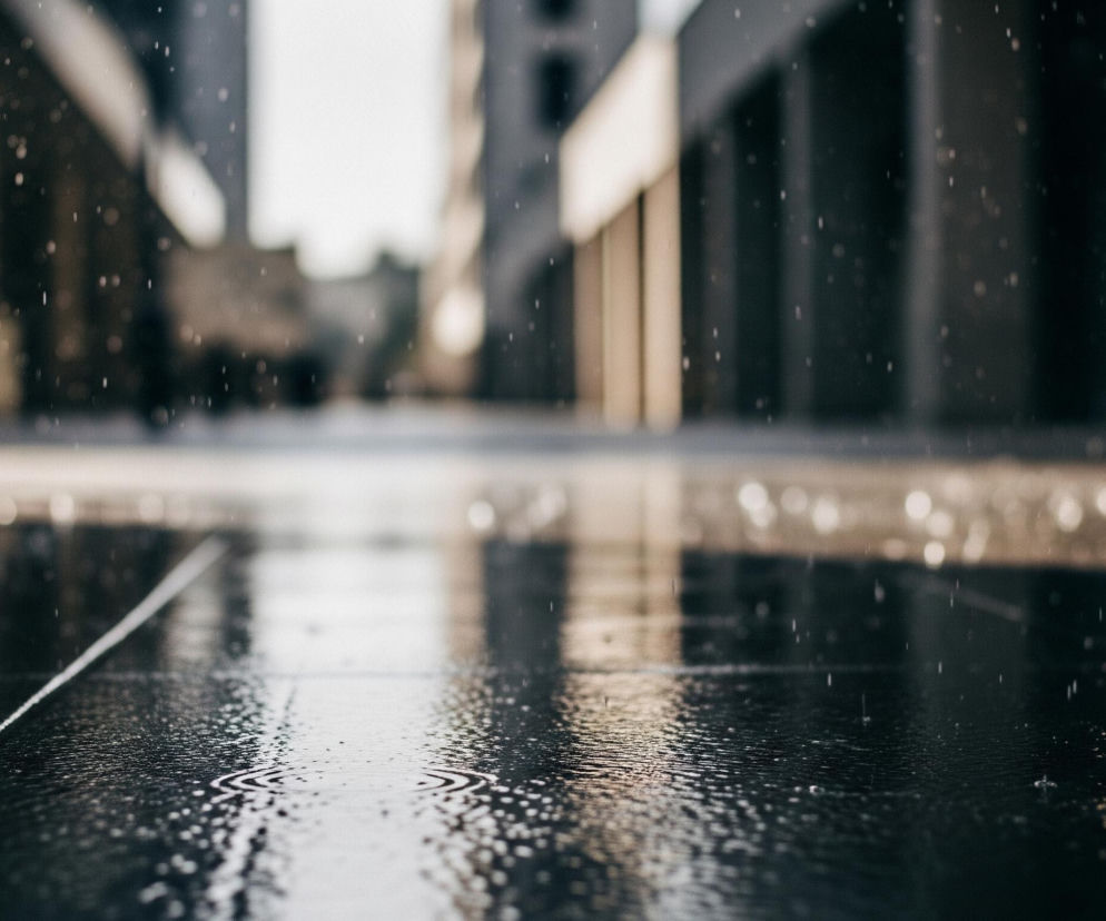 Pierwszy plan przedstawia mokrą, ciemną, kamienną posadzkę, na której widoczne są liczne krople wody oraz drobne, koncentryczne fale na powierzchni. Pozioma fuga dzieli posadzkę, a w oddali, w rozmazanym tle, widać sylwetki budynków o prostej architekturze. Ponadto, w kadrze widoczne są jasne, rozmyte punkty światła, wyglądające jak spadające krople deszczu.