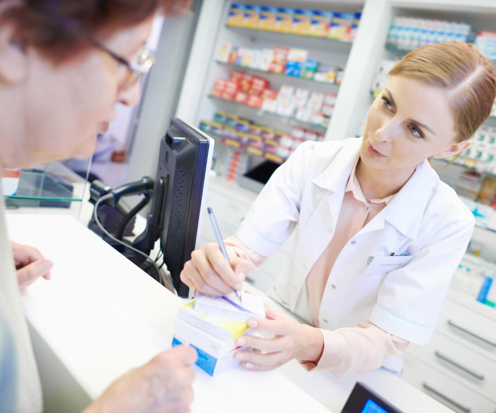 Farmaceutka w białym fartuchu rozmawia ze starszą kobietą, która stoi przy ladzie w aptece. Farmaceutka trzyma długopis i patrzy na klientkę, doradzając w wyborze leków. Więcej informacji o działaniu płynu Lugola znajdziesz na Poradnik Zdrowie.