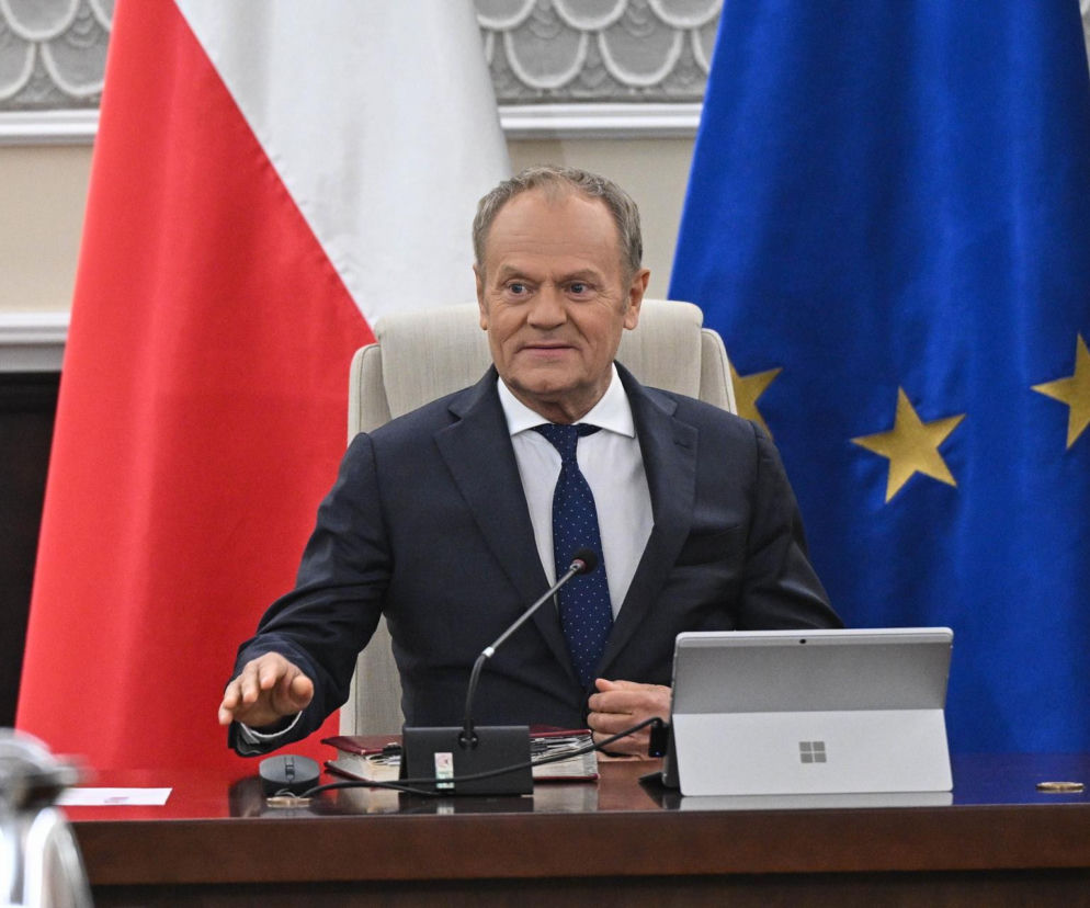 Premier Donald Tusk siedzi za stołem podczas nadzwyczajnego posiedzenia rządu, na tle flag Polski i Unii Europejskiej, omawiając radykalne obniżki VAT i akcyzy na paliwa, o czym przeczytasz więcej na Super Biznes.