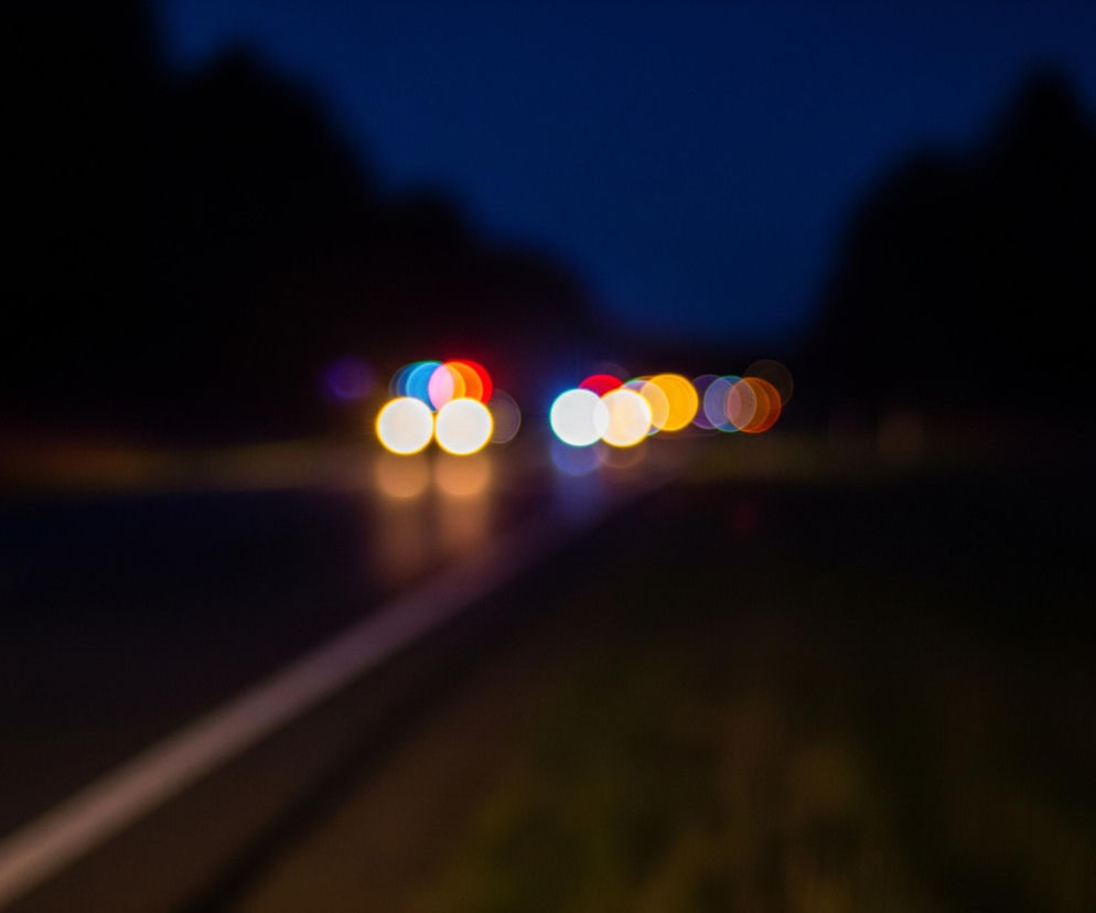 Rozmyty obraz przedstawia nocną scenę drogową z widocznymi światłami pojazdów. W centralnej części kadru znajdują się rzędy okrągłych, jaskrawych świateł w odcieniach białego, czerwonego, niebieskiego i pomarańczowego, odbijające się delikatnie na ciemnej nawierzchni drogi. Biała, ciągła linia wyznacza prawy skraj drogi na pierwszym planie, biegnąca ukośnie od lewego dolnego rogu ku środkowi kadru. Tło stanowi ciemne niebo w górnej części oraz niewyraźne, ciemne sylwetki drzew lub wzniesień po bokach drogi.