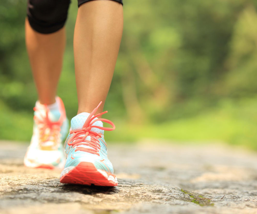 Nogi kobiety w sportowych butach z pomarańczowymi sznurówkami i turkusowymi wstawkami, poruszającej się po kamienistej ścieżce, z niewyraźnym, zielonym tłem lasu. Więcej o wyzwaniach spacerowych przeczytasz na naszym portalu.