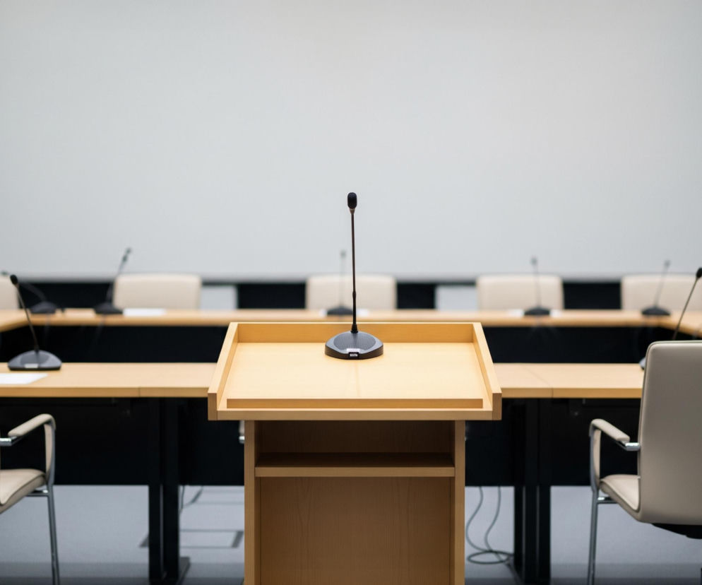 Na pierwszym planie, centralnie, stoi drewniana mównica w jasnym odcieniu brązu, z płaskim blatem i niewielkimi półkami poniżej. Na blacie mównicy znajduje się czarny mikrofon na cienkiej, elastycznej nóżce, skierowany ku górze. Za mównicą rozciąga się duży, drewniany stół konferencyjny w kształcie litery U lub owalu, wokół którego rozmieszczone są liczne, jasnobrązowe krzesła z metalowymi ramami i podłokietnikami, skierowane przodem do mównicy. Na stole, przed każdym miejscem siedzącym, widoczne są małe, czarne mikrofony. W tle znajduje się duży, biały ekran projekcyjny, zajmujący całą szerokość ściany. Cała scena utrzymana jest w stonowanych barwach beżu, brązu, szarości i czerni.