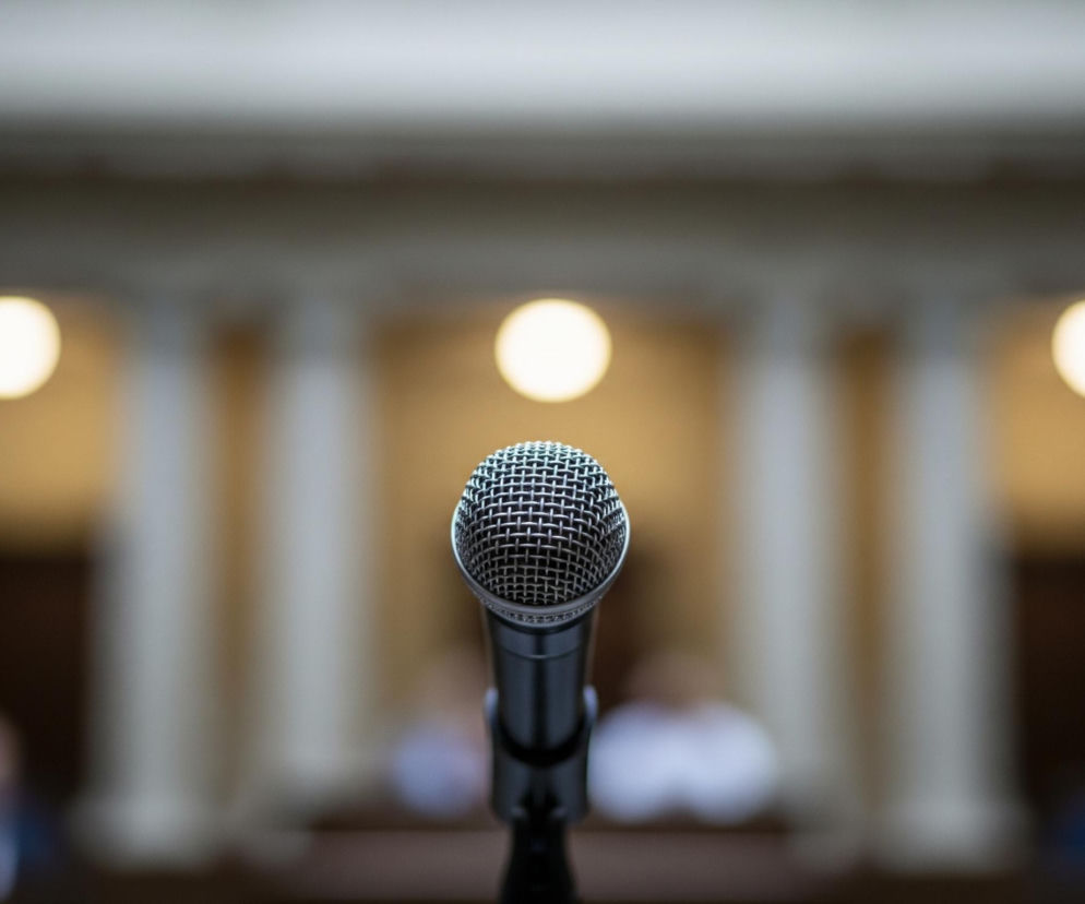 Centralnie na pierwszym planie znajduje się mikrofon z metalową, siatkową główką, mocno zogniskowany i wyraźny. Mikrofon jest czarny i srebrny, a jego konstrukcja jest widoczna w drobnych detalach. Tło jest rozmyte, ale widać na nim elementy architektury, takie jak jasne filary lub kolumny oraz trzy okrągłe, rozświetlone punkty, które wyglądają jak lampy lub oprawy oświetleniowe, emitujące ciepłe, żółtawe światło. Całość ma stonowane barwy, z dominującymi szarościami, beżami i brązami w tle oraz intensywną czernią i srebrem mikrofonu.