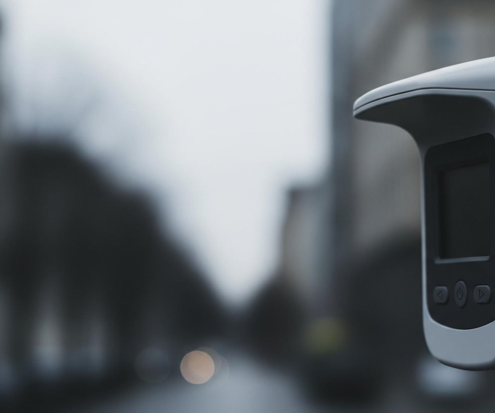 Szare urządzenie z wyświetlaczem i trzema przyciskami widać po prawej stronie kadru. Tło jest rozmyte, ukazując szare, wysokie budynki i pasmo jasnego, zachmurzonego nieba. Na dole, w oddali, dostrzegalne są dwie rozmyte plamy światła, jedna żółtawa i jedna bledsza.