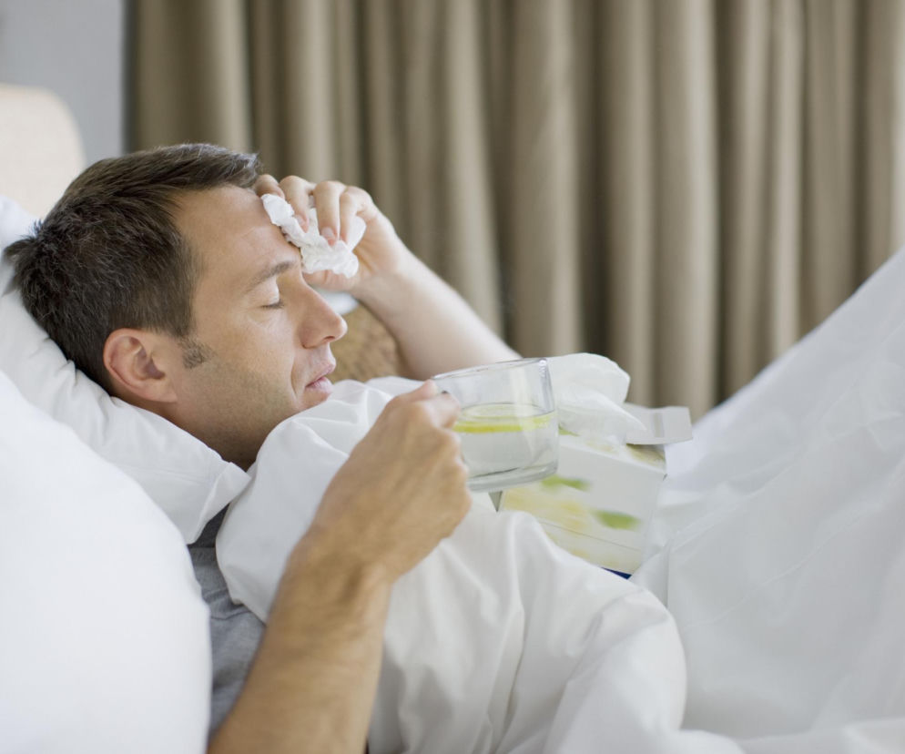 Mężczyzna w łóżku z zamkniętymi oczami, z okładem na czole i szklanką z wodą z cytryną. Obok leżą chusteczki higieniczne, co wskazuje na grypę lub przeziębienie. Na Poradnik Zdrowie znajdziesz informacje o powrocie COVID i supergrypy, objawach i profilaktyce.