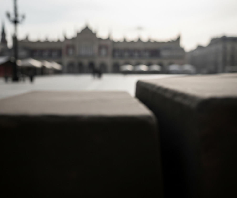 Na pierwszym planie, w dolnej części obrazu, widoczne są dwa ciemne, rozmyte, prostokątne obiekty o zaokrąglonych krawędziach, sprawiające wrażenie kamiennych ław. Jeden obiekt znajduje się bliżej lewej krawędzi, drugi nieco dalej, po prawej stronie. Za nimi rozciąga się przestronny, jasnoszary plac. W tle, na środku kadru, rozmyty jest duży, jasno-szary budynek o wyraźnych elementach architektonicznych, takich jak arkady i zdobione szczyty dachu. Po lewej stronie placu widać również rozmyte budynki i ciemną latarnię uliczną, a po prawej stronie placu inne rozmyte budowle. Cały obraz utrzymany jest w stonowanej, niemal monochromatycznej palecie barw, głównie szarości i beże, z kontrastującymi ciemnymi elementami na pierwszym planie.