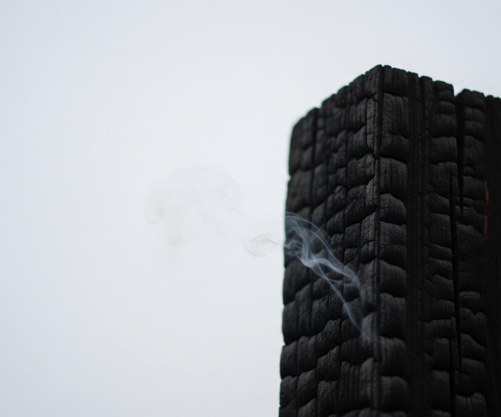 Duży, prostokątny, zwęglony kawałek drewna o głębokiej czarnej barwie zajmuje prawą stronę kadru, z jego wyraźną, pofałdowaną strukturą powierzchni. Z lewej krawędzi drewna unosi się smuga delikatnego, białego dymu, który rozprasza się w kierunku lewej części obrazu. Tło jest jednolicie jasnoszare, co kontrastuje z ciemnym drewnem i podkreśla ulotność dymu.