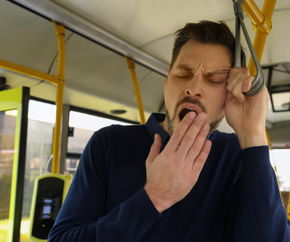 Mężczyzna w niebieskiej koszulce ziewający i zakrywający usta dłonią podczas podróży autobusem, sugerujący zmęczenie lub senność. Na portalu Poradnik Zdrowie znajdziesz informacje o przyczynach i skutkach nadmiernego ziewania oraz o tym, co może sygnalizować.