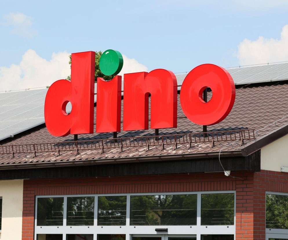 Czerwony neonowy napis Dino na tle dachu z panelami fotowoltaicznymi symbolizuje sieć handlową w kontekście kontrowersji wokół warunków pracy, o których można przeczytać na Super Biznes.