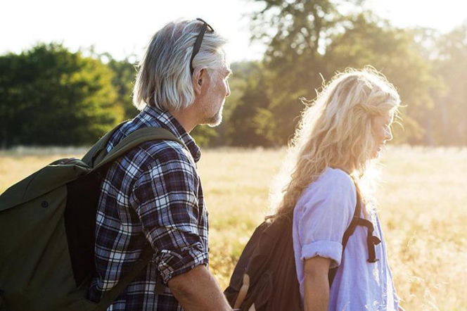 Starszy mężczyzna w kraciastej koszuli i młodsza kobieta o blond włosach spacerują po łące, mając na plecach plecaki, symbolizując aktywność fizyczną mimo nietrzymania moczu, o czym więcej przeczytasz na Poradnik Zdrowie.