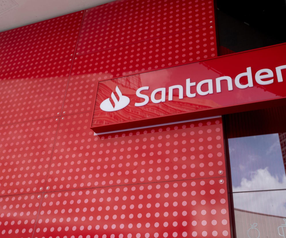 Czerwony szyld banku Santander z białym logo i napisem, widoczny na tle czerwonej ściany z okrągłymi wzorami, ilustrujący rebranding na Erste Bank Polska. Więcej informacji o zmianach w bankowości przeczytasz na Super Biznes.