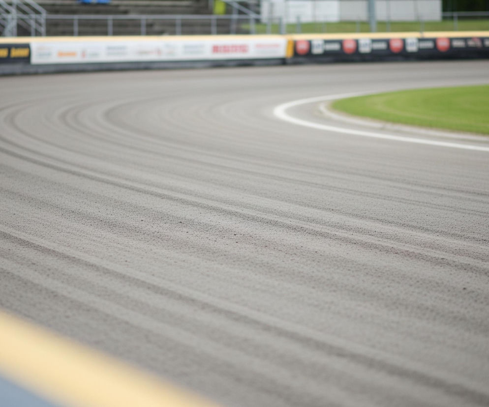 Na pierwszym planie widoczny jest fragment toru żużlowego o piaszczystej, szarej nawierzchni, z wyraźnymi równoległymi śladami, biegnący w lekkim łuku od dolnego lewego rogu w stronę środka kadru. W dolnym lewym rogu toru znajduje się szary element, a na jego krawędzi widoczny jest żółty pas. Tor od prawego boku jest ograniczony cienką, białą linią, za którą rozciąga się zielony pas trawy.

W tle, za pasem trawy, znajduje się biało-czarna banda reklamowa z niewyraźnymi napisami i logotypami, biegnąca wzdłuż zakrętu toru. Ponad bandą widać elementy trybun sportowych, metalowe barierki oraz fragment zieleni drzew i jasnego, pochmurnego nieba. Całość tworzy scenę stadionu sportowego, prawdopodobnie żużlowego, z pustymi trybunami, co sugeruje, że zdjęcie zostało wykonane poza czasem zawodów.
