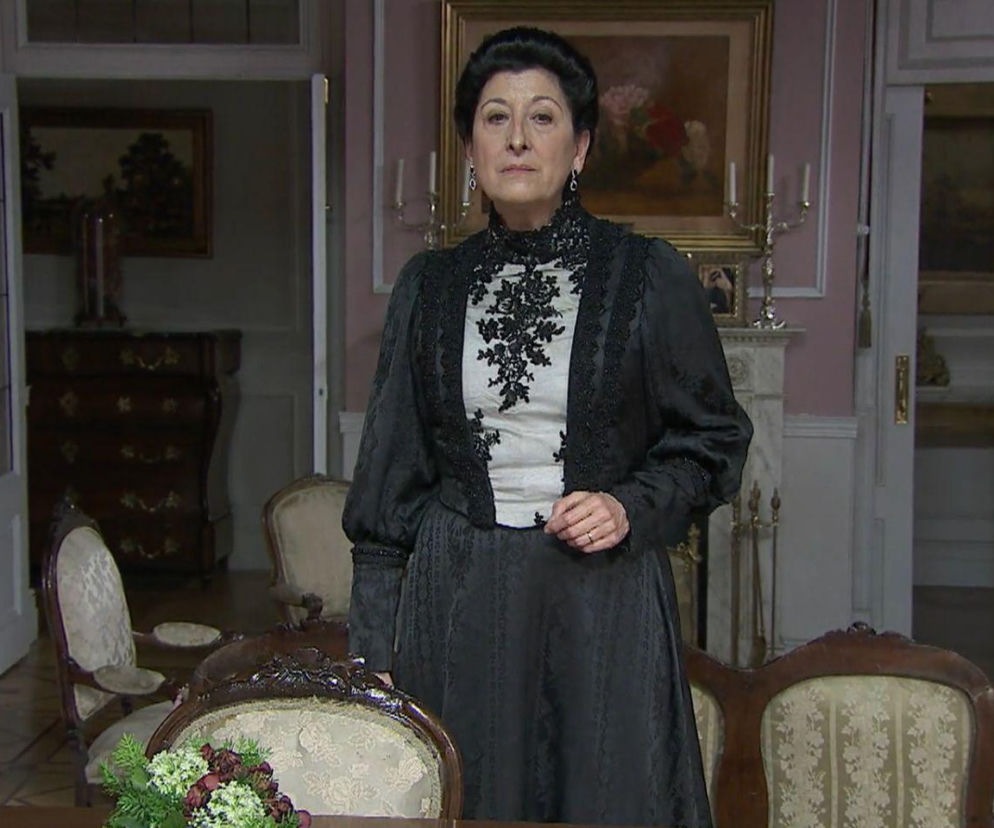 Ucharakteryzowana aktorka z serialu Akacjowa 38 w ciemnym stroju z koronkowymi zdobieniami na bluzce, stoi w eleganckim salonie z kominkiem i obrazami. To postać z popularnego serialu, o którym przeczytasz więcej na naszym portalu.