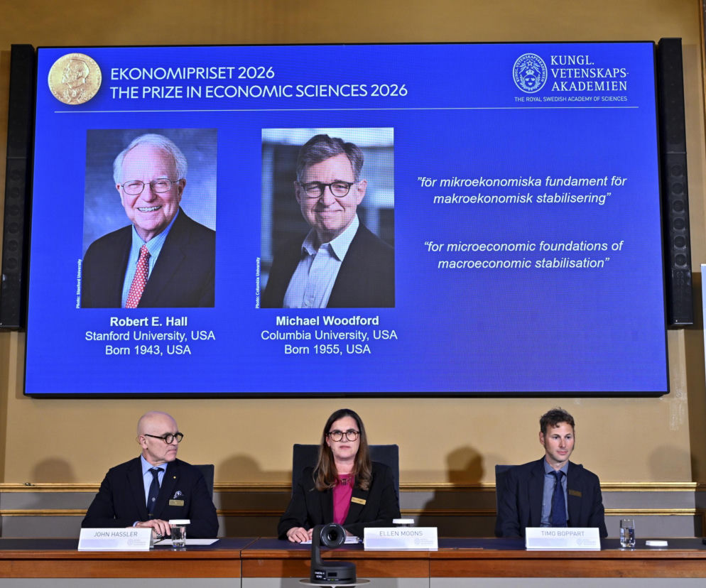 Troje członków Akademii Nauk za stołem. Na ekranie zdjęcia laureatów Roberta Halla i Michaela Woodforda w SE Superbiz.