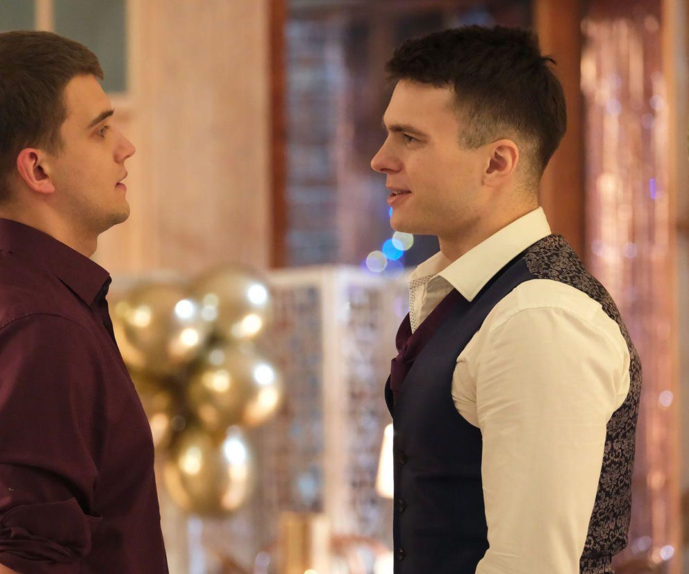 Mateusz (Rafał Kowalski) i Tomek Kubicki (Chris Cugowski) stojący naprzeciwko siebie podczas kłótni na weselu w serialu M jak miłość. Mateusz, ubrany w bordową koszulę, patrzy na Tomka, który w białej koszuli i kamizelce z wzorami, uśmiecha się. W tle złote balony i dekoracje imprezowe. Więcej o tym na Super Seriale.