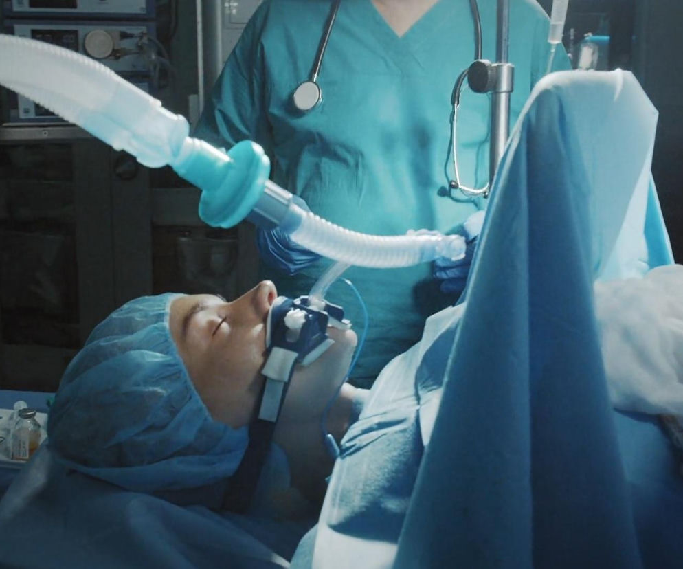 Pacjentka Dominika Kachlik jako Franka leży na stole operacyjnym w czepku, z maską tlenową na twarzy, podłączona do aparatury medycznej. Obok stoi anestezjolog w zielonym stroju, obserwując parametry. Scena z M jak miłość przedstawia dramatyczne chwile śmierci bohaterki, o których możesz przeczytać więcej na Super Seriale.