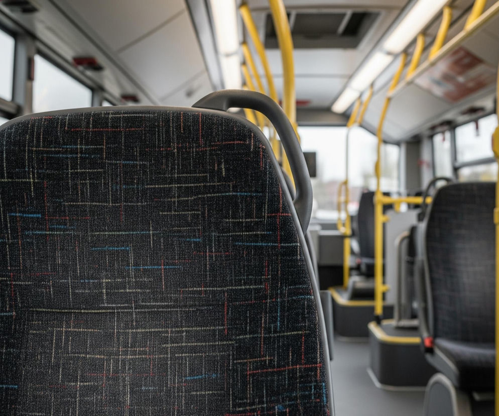 Wnętrze autobusu widoczne jest z perspektywy siedzącego pasażera, z dużym zbliżeniem na oparcie przedniego siedzenia. Oparcie to, wykonane z ciemnoszarej, prawdopodobnie tekstylnej tapicerki, pokryte jest wzorem drobnych, przecinających się linii w kolorach czerwonym, niebieskim, żółtym i białym. Nad siedzeniem widoczny jest szary uchwyt do trzymania. W tle rozciąga się pusta przestrzeń autobusu z rzędami identycznych siedzeń, żółtymi rurkami do trzymania, podłużnymi lampami sufitowymi oraz dużymi oknami, przez które widać niewyraźny krajobraz.