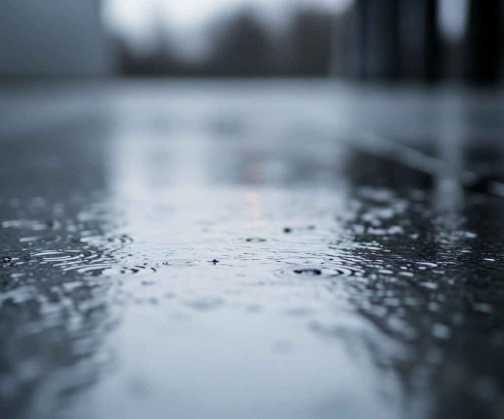 Pierwszy plan przedstawia mokrą, błyszczącą nawierzchnię z licznymi kroplami deszczu i małymi kręgami na wodzie, co sugeruje opady. Powierzchnia, będąca w centrum uwagi, jest szaro-niebieska, z jaśniejszymi odbiciami. W tle widoczne są rozmyte kontury budynków i drzew, z dominującymi ciemnymi odcieniami szarości i błękitu, tworząc wrażenie pochmurnego dnia.