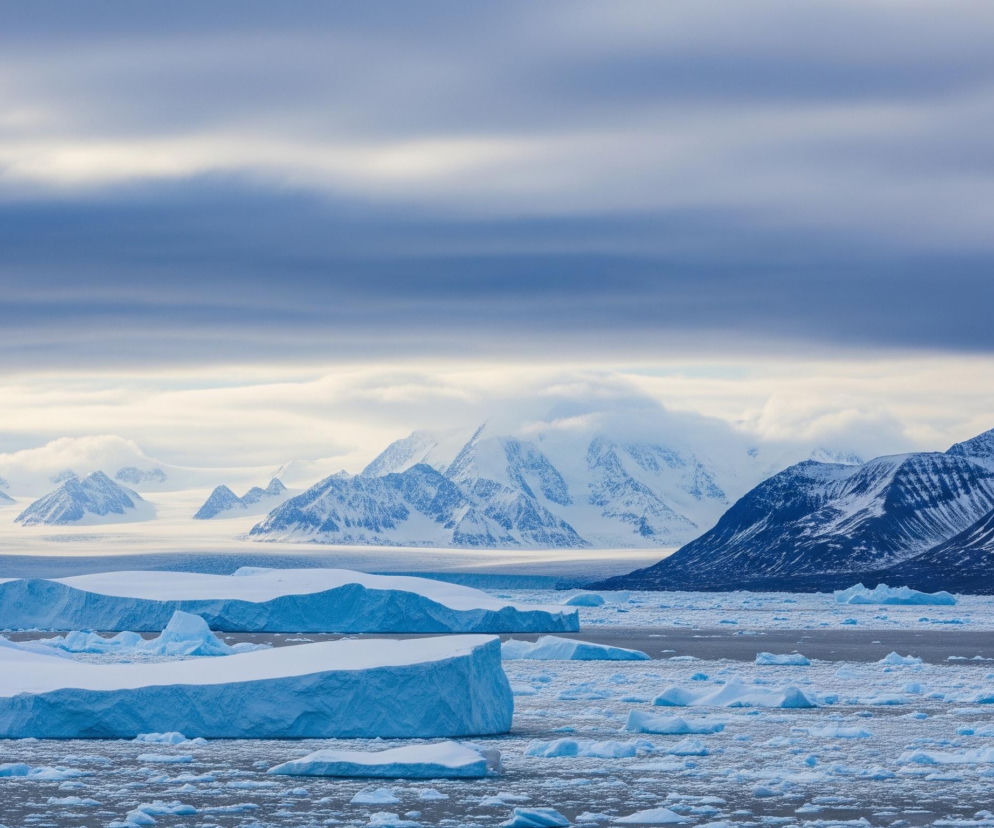 Rozległy, zimowy krajobraz przedstawia zamarznięty akwen z licznymi górami lodowymi i mniejszymi kawałkami lodu na pierwszym planie, w jasnych odcieniach błękitu i bieli. Na horyzoncie widoczne są ośnieżone, skaliste góry, z częściowo zachmurzonym niebem w tle, gdzie dominują szare i jasne obłoki. Centralnie umiejscowione są dwa duże błękitne bloki lodu, a pomiędzy nimi rozciąga się pasmo górskie częściowo zasłonięte przez białe chmury.