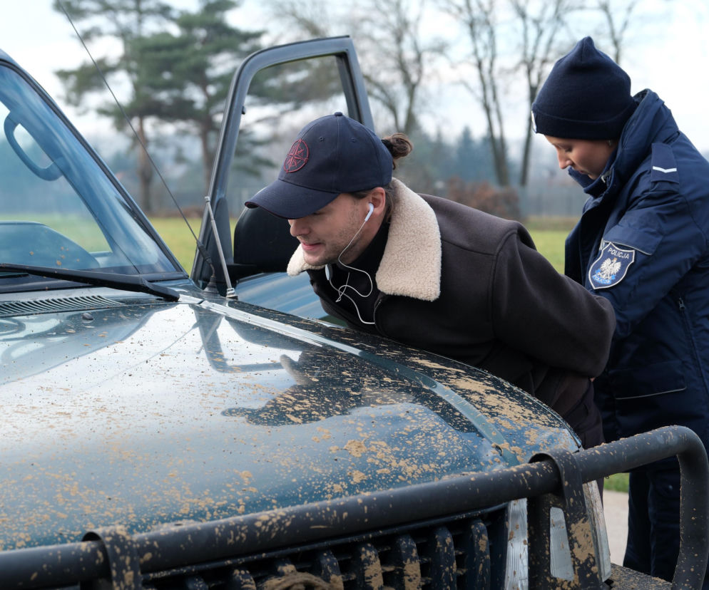 Natalka Mostowiak (Dominika Suchecka) w mundurze policjantki zakłada kajdanki Mikołajowi Lipińskiemu (Mateusz Kościukiewicz), który opiera się o brudną, terenową maskę samochodu. Scena z serialu M jak miłość, odcinek 1927. Więcej na Super Seriale.