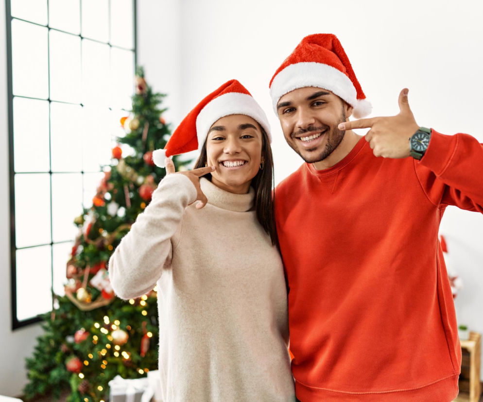 Uśmiechnięta para w czapkach Mikołaja, wskazująca palcami na swoje białe zęby. Stoją na tle choinki, symbolizując zdrowy uśmiech jako świąteczny prezent. Na portalu Poradnik Zdrowie znajdziesz wskazówki, jak dbać o piękny i zdrowy uśmiech.