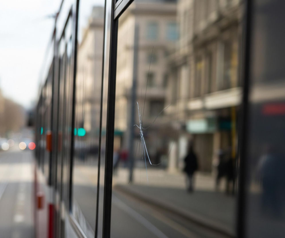 Duża szyba tramwaju z widocznym pęknięciem w kształcie gwiazdy dominuje prawą stronę obrazu, oddzielając widok z wnętrza pojazdu od zewnętrznego krajobrazu miejskiego. Przez szybę, a także po lewej stronie kadru, rozciąga się ulica z widocznymi torami tramwajowymi, budynkami po obu stronach i rozmazanymi sylwetkami pieszych oraz świateł sygnalizacyjnych w kolorach zielonym i czerwonym. Odbicia budynków i światła ulicznego są widoczne na powierzchni szyby, szczególnie po prawej stronie, co tworzy wrażenie głębi i złożoności miejskiego otoczenia.