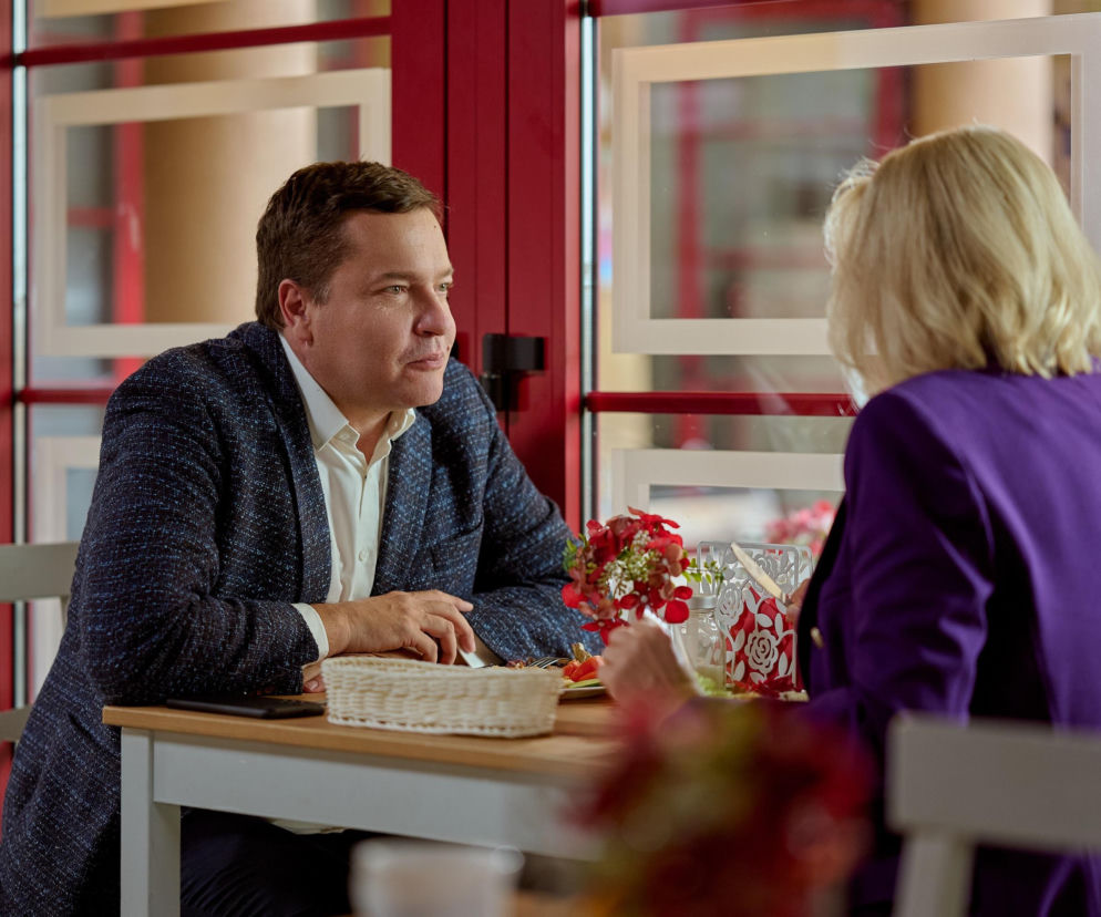 Michał z serialu „Na Wspólnej” w granatowej marynarce i białej koszuli, siedzi przy stole z bukietem kwiatów i rozmawia z kobietą o blond włosach, ubraną w fioletowy żakiet. To zdjęcie z planu serialu „Na Wspólnej”, a więcej o nim przeczytasz na Super Seriale.