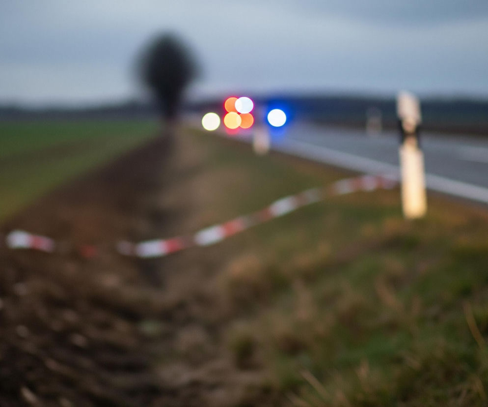 Na pierwszym planie, w lewym dolnym rogu, znajduje się rozmyte pole z brązową, zaoraną ziemią, a obok niego, po prawej stronie, pas zielonej trawy. Przez środek obrazu, w poziomie, przebiega biało-czerwona taśma, która oddziela pole od pasa zieleni i prowadzi w głąb kadru. W tle, po prawej stronie, widać rozmazaną drogę z białą linią, a dalej na horyzoncie, pojedyncze drzewo i grupę świateł – czerwonych, pomarańczowych i niebieskich, wskazujących na pojazdy uprzywilejowane. Niebo jest pochmurne i szare, a cały obraz jest nieostry, z efektem bokeh, co skupia uwagę na taśmie i światłach w oddali.