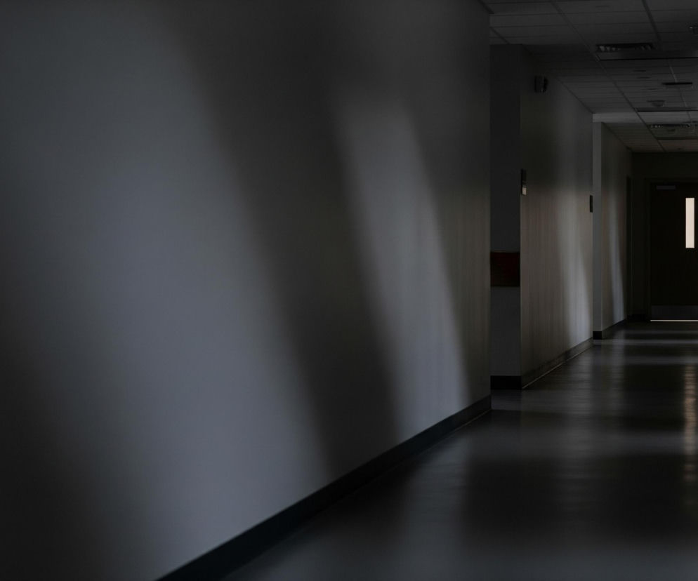 Ciemny korytarz z kremowymi ścianami rozciąga się w głąb obrazu, z lewą ścianą blisko kadru i ciemniejszą. Na końcu korytarza widać otwarte drzwi, za którymi widać jasne, białe światło. Podłoga w korytarzu jest ciemna i lśniąca, odbijając światło padające z dalszych partii. Sufit jest kasetonowy, również w ciemnych odcieniach, z widocznymi elementami oświetlenia liniowego.