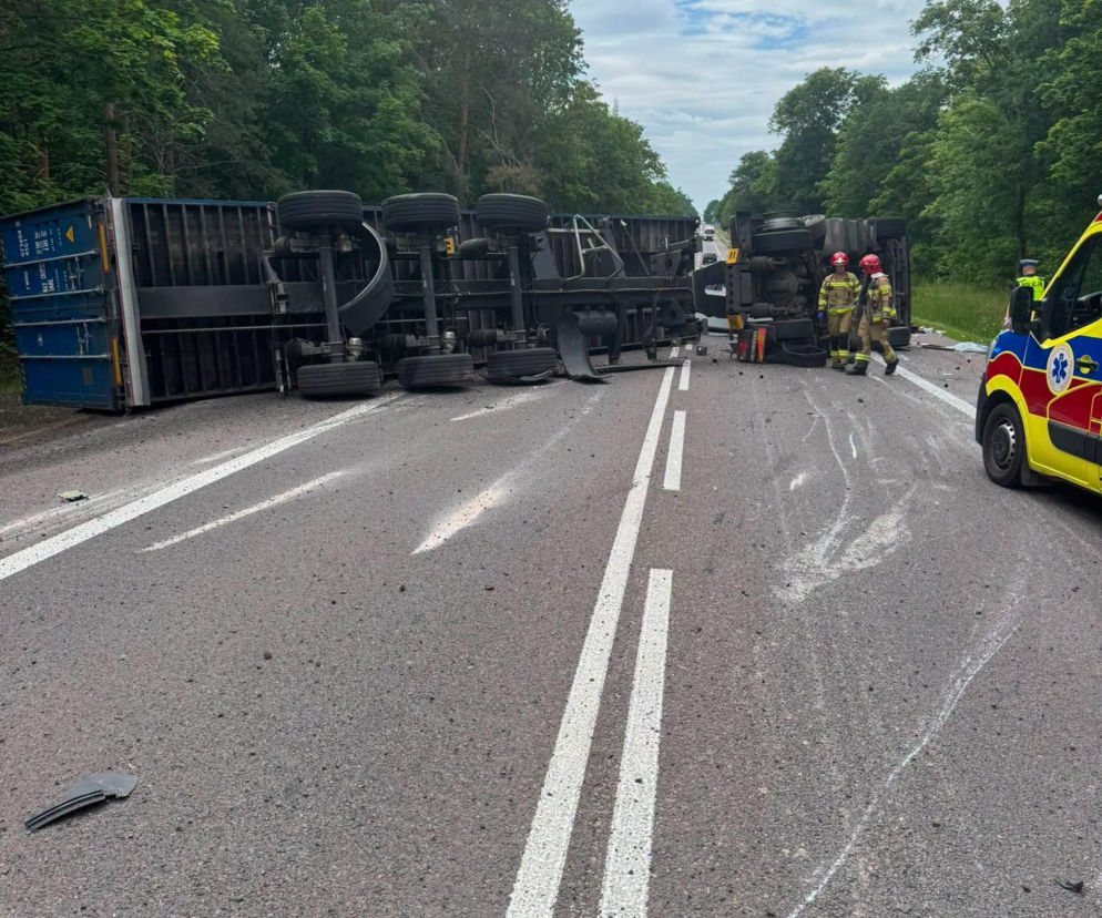 Przewrócona na bok ciężarówka z niebieskim kontenerem blokuje oba pasy drogi krajowej w Worońcu. Obok wraku pojazdu stoją strażacy w pełnym umundurowaniu, a nieopodal widoczny jest bok karetki. W tle widać las, a nad nim zachmurzone niebo. Więcej informacji o zdarzeniu znajdziesz na naszym portalu.