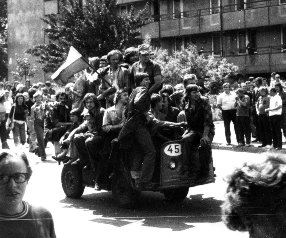 Młodzi ludzie w trakcie protestu robotniczego w Radomiu w czerwcu 1976 roku. Część protestujących jedzie na trójkołowym pojeździe z numerem 45, a jeden z nich trzyma flagę narodową. W tle widoczny tłum i budynki. Więcej o 50 rocznicy protestu na naszym portalu.