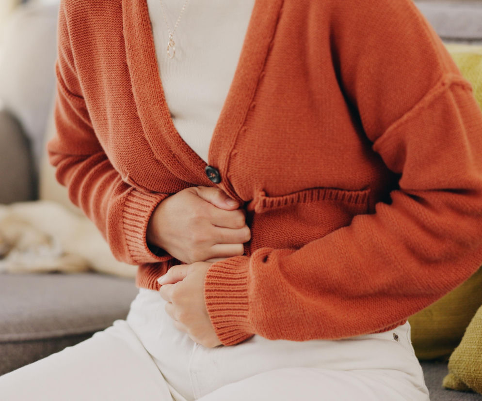 Kobieta w pomarańczowym kardiganie i białych spodniach siedząca na sofie, trzymająca się za brzuch, z miną wskazującą na ból. Zdjęcie ilustruje dolegliwości jelitowe, o których piszemy na portalu Poradnik Zdrowie.