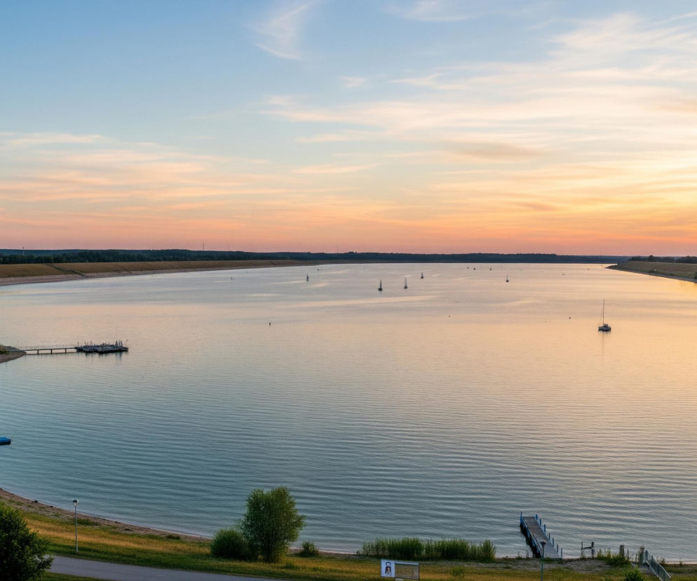 Rozległy, spokojny zbiornik wodny zajmuje centralną część obrazu, otoczony po obu stronach zielonymi, trawiastymi brzegami, które łagodnie wznoszą się w kierunku lasów widocznych na horyzoncie. Na błękitno-pomarańczowym niebie, widocznym w górnej części kadru, rozciągają się delikatne, poziome chmury, a ich barwy odbijają się w lustrzanej powierzchni wody. Po lewej stronie, wzdłuż zakrzywionego brzegu, prowadzi asfaltowa droga, przy której rosną drzewa i krzewy, a także znajduje się mały pomost i zacumowane łodzie; po prawej stronie, również wzdłuż brzegu, widać kolejny pomost i pojedynczą łódź. Na wodzie unosi się kilka białych żaglówek oraz motorówek, rozproszonych po całym akwenie.