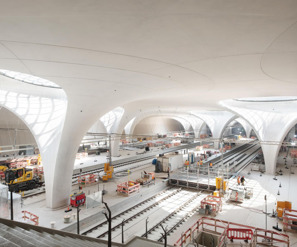 Wnętrze w budowie podziemnego dworca Stuttgart Hauptbahnhof z widocznymi betonowymi filarami w kształcie kielichów, świetlikami na dachu oraz torami kolejowymi i pracownikami. O innowacyjnym projekcie architektonicznym przeczytasz na Architektura Murator Plus.